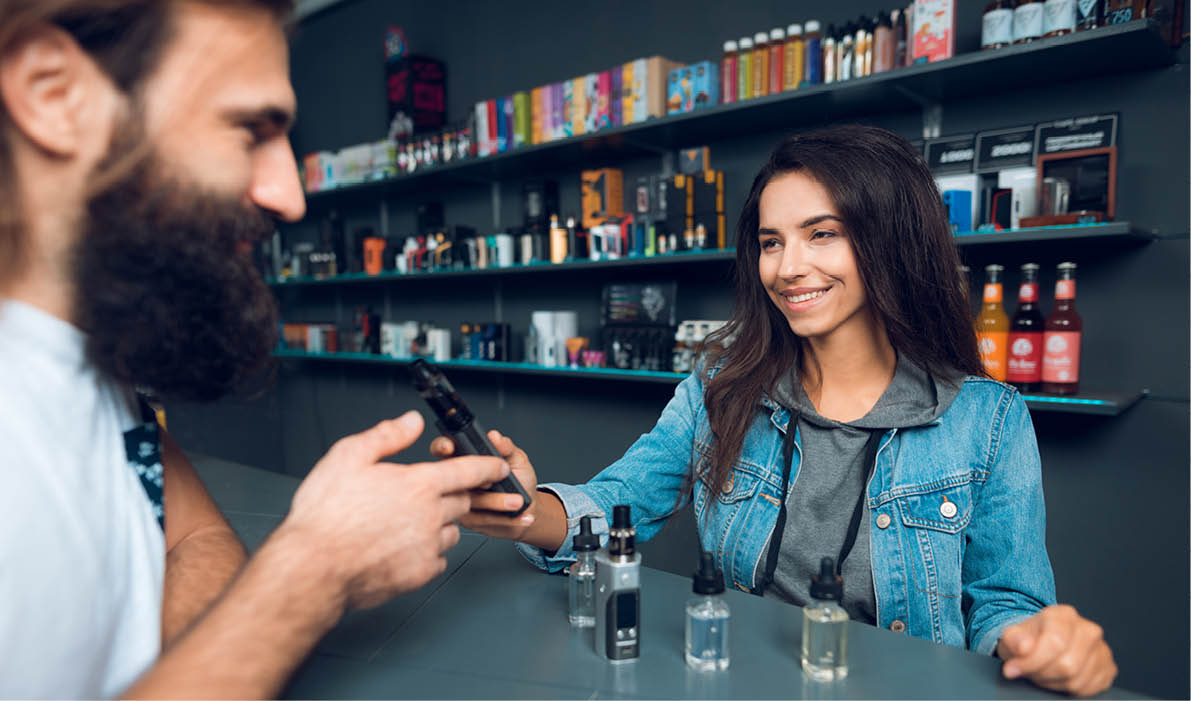 Girl seller shows the choice of electronic cigarettes in vapeshop. Nearby is a buyer - a man with a beard. The store has a large assortment of electronic cigarettes.