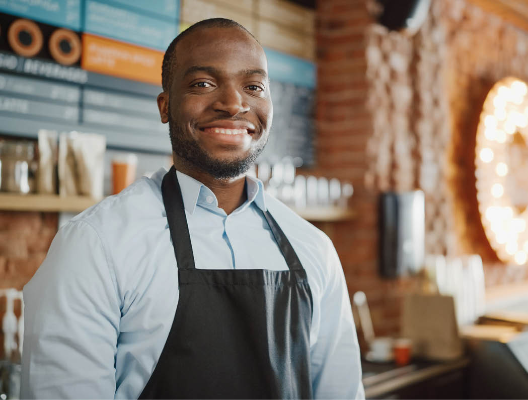 Handsome Black African American Barista with Short Hair and Beard Wearing Apron is Smiling in Coffee Shop Restaurant  Portrait of Happy Employee Behind Cozy Loft-Style Cafe Counter 