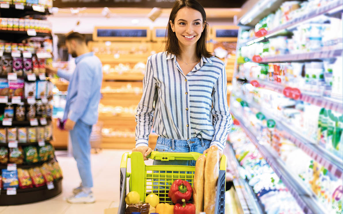 Smiling Cheerful Young Woman Walking With Shopping Trolley Cart Along The Shelves In Grocery Store  Happy Female Customer Buying Groceries In Supermarket, Looking At Fridge With Dairy Products