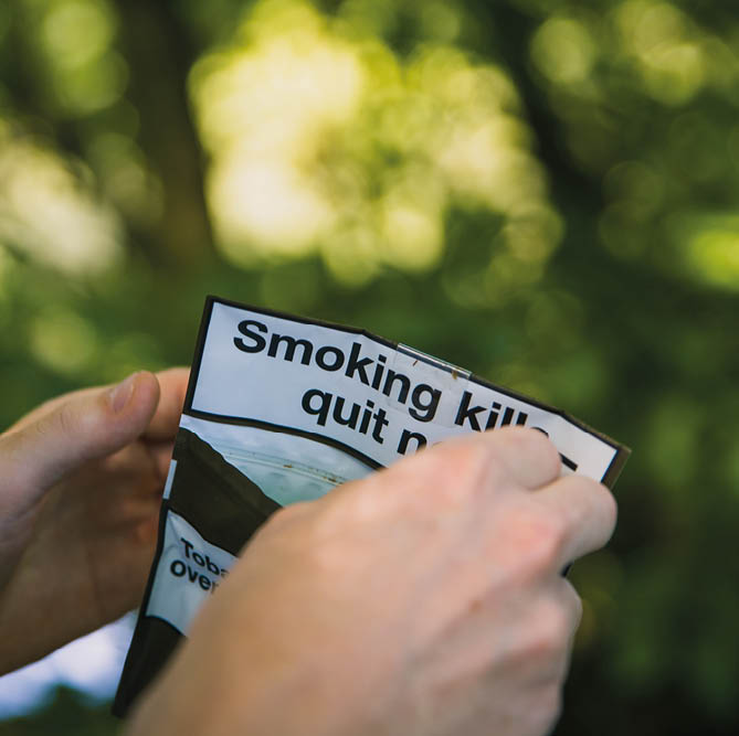 Thornton, England, 24 08 19 Man under the influence of drugs or alcohol struggling to roll a tobacco cigarette or marijuana joint or spliff in the wood forest with nice sunny green bokeh background 