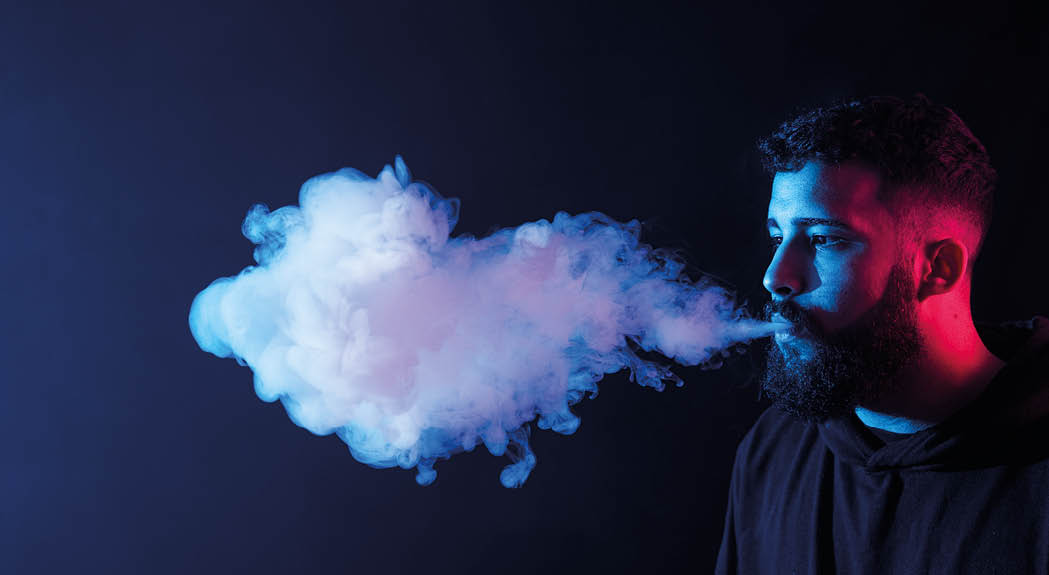 Young bearded arab man smoking vape and doing tricks in dark studio with colored lights  Copy space 