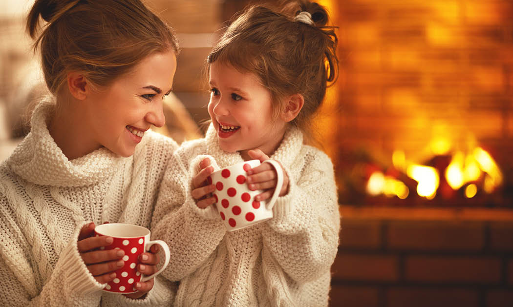 family mother and child daughter drinking tea and laughing on winter evening by fireplace