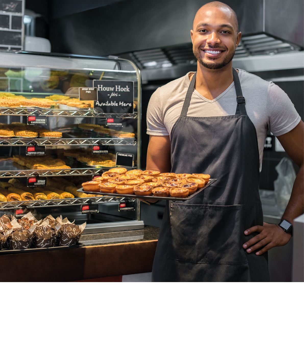 Smiling black baker with apron holding tray of small pastry and looking at camera. Young african chef holding sweet tray at cafeteria. Happy black man smiling at bakery.