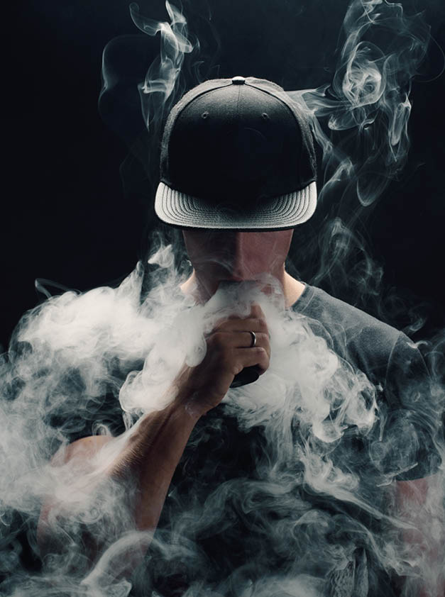 Vape man. Portrait of a handsome young white guy in a modern black cap vaping and letting off puffs of steam from an electronic cigarette 