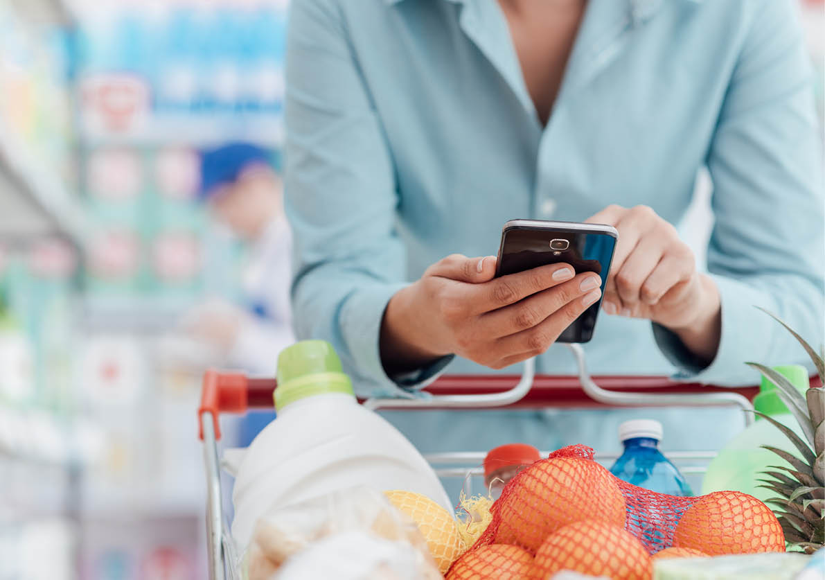 Woman doing grocery shopping at the supermarket, she is leaning on the shopping cart and connecting with her phone, apps and retail concept