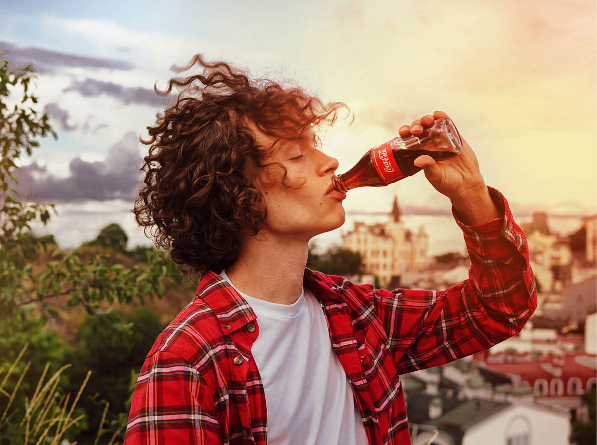 Kyiv, Ukraine, August 1, 2020  Handsome young man with curly hair and in a red plaid shirt holds a glass bottle of Coca-Cola in his hand and drinks from it 