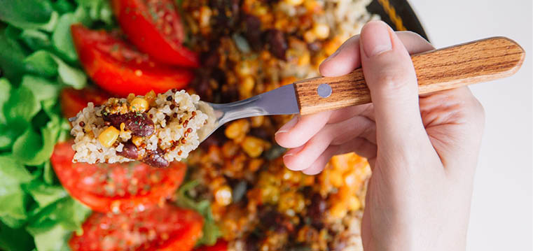 Top view of Female hands at dinner table holding fork above plate with quinoa, red beans, corn, tomato and green salad, close up