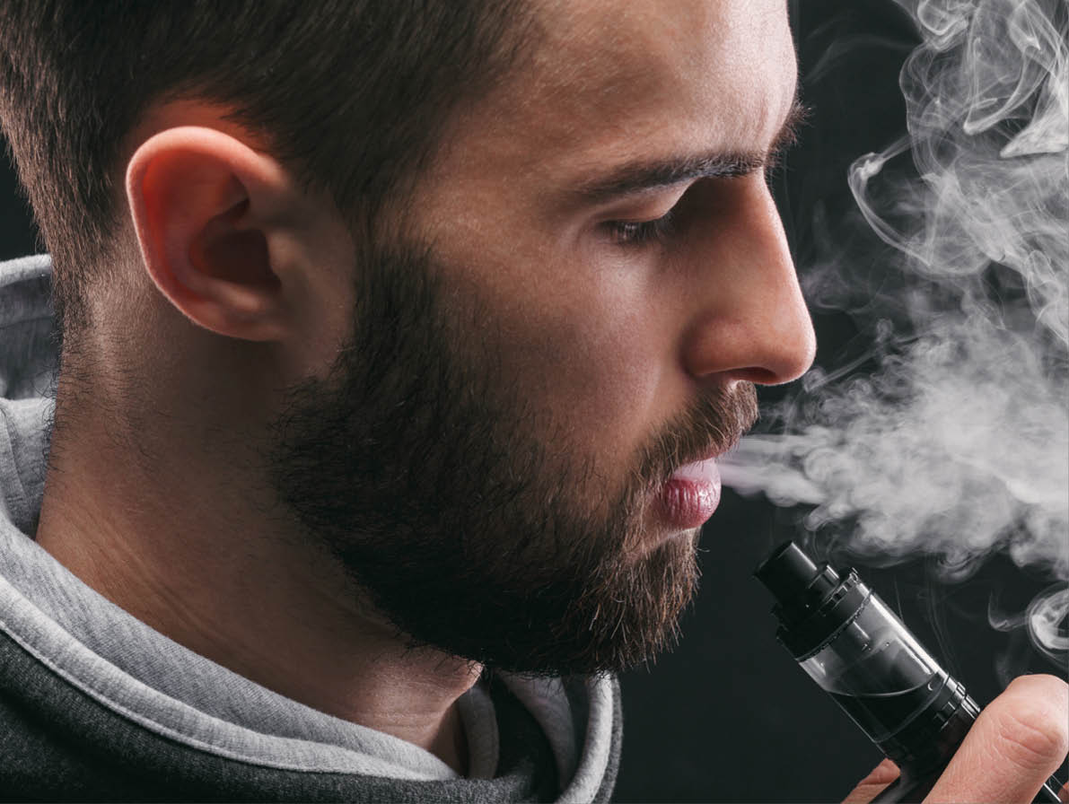 Man with vaping mod exhaling steam at black studio background. Bearded guy smoking e-cigarette to quit tobacco. Vapor and alternative nicotine free smoking concept, closeup