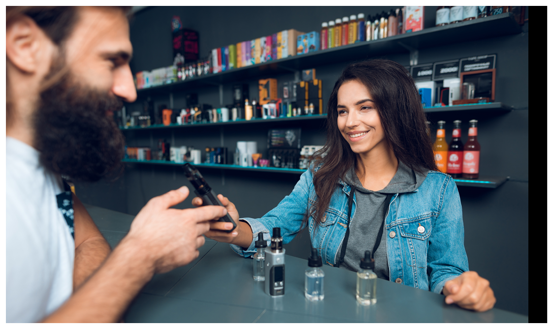 Girl seller shows the choice of electronic cigarettes in vapeshop. Nearby is a buyer - a man with a beard. The store has a large assortment of electronic cigarettes.