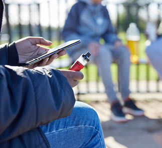 Close Up Of Teenagers With Mobile Phone Vaping and Drinking Alcohol In Park