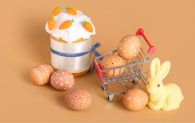 Small shopping cart with Easter cake, eggs and bunny figurine on beige background