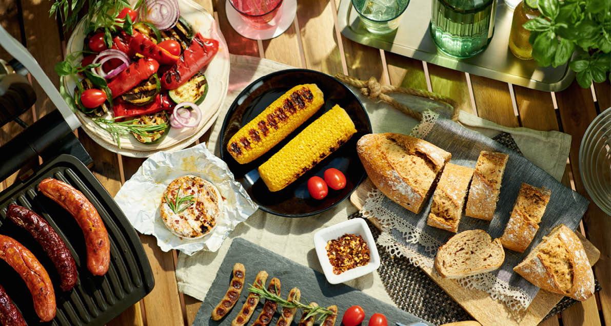 Summer backyard party. Upper view of desk with electric grill, grilled sausages, vegetables, corn, bread and camembert.