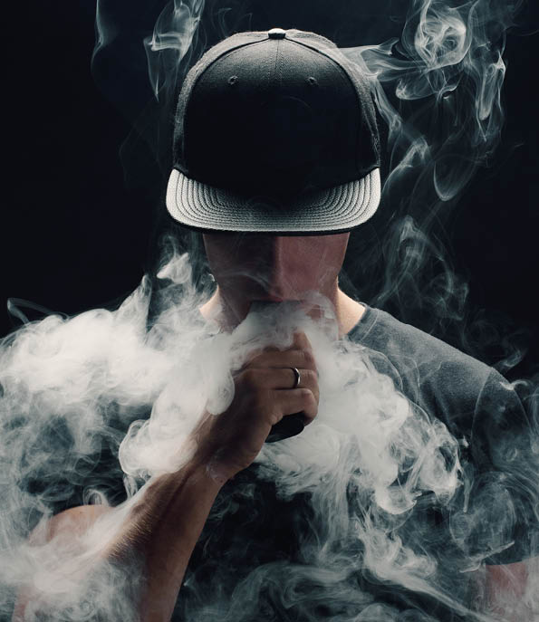 Vape man. Portrait of a handsome young white guy in a modern black cap vaping and letting off puffs of steam from an electronic cigarette 
