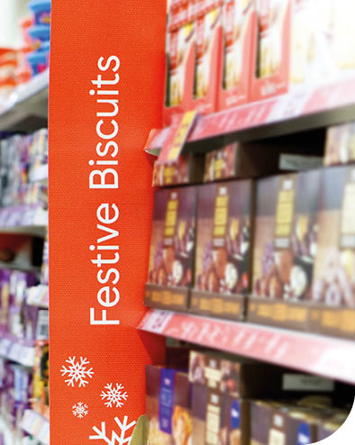 Christmas shoppers in a supermarket aisle with focus on a Festive Biscuits sign.