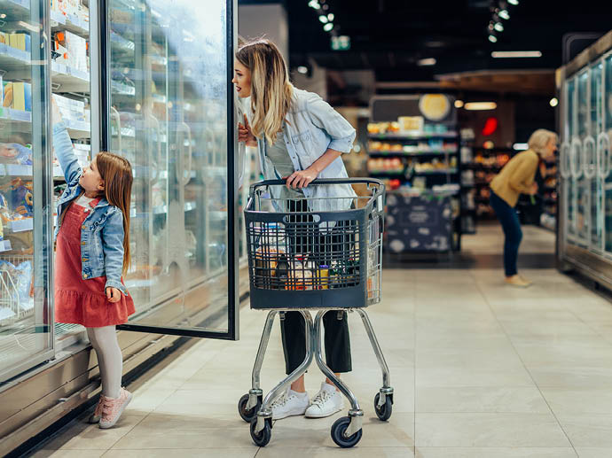 Young beautiful mother holding pushing shopping cart with her child in supermarket. Girl is choosing daily milk product picking up from shelf with her mother beside. Shopping for healthy.