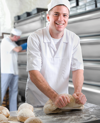A medium shot of a young baker smiling and looking the camera while kneading bread dough in a bakery.