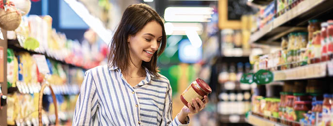Portrait Of Smiling Woman With Shopping Cart In Supermarket Buying Groceries Food Walking Along The Aisle And Shelves In Grocery Store, Holding Glass Jar Of Sauce, Choosing Healthy Products In Mall