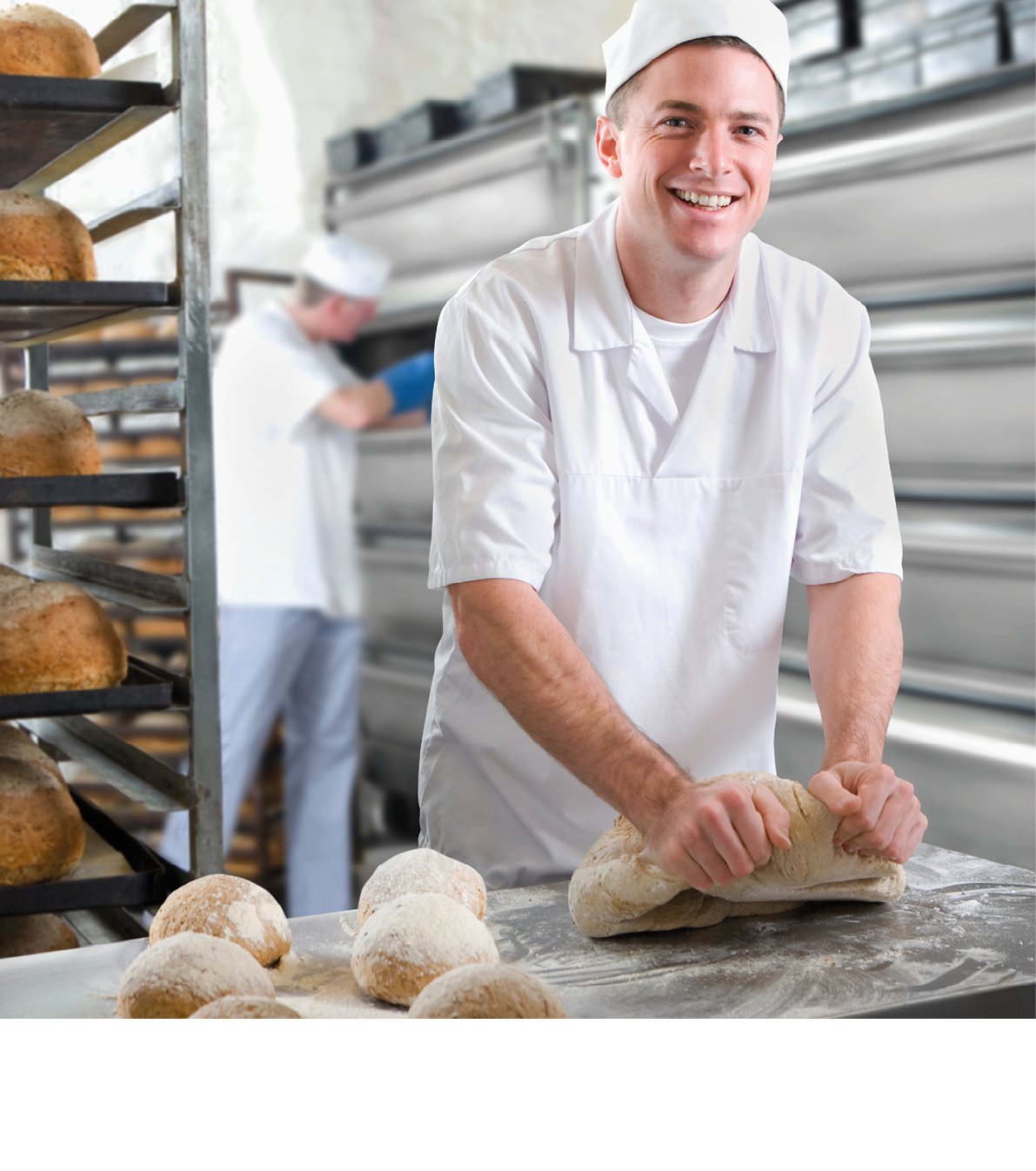A medium shot of a young baker smiling and looking the camera while kneading bread dough in a bakery.