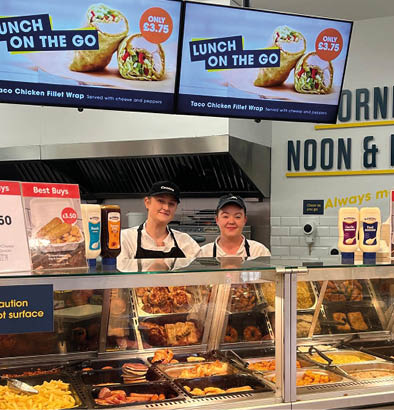 Two people are standing behind a deli counter with a variety of food items, including sandwiches and hot dogs. AI generated content