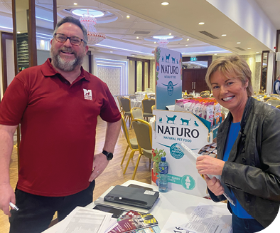 A man and woman are standing in front of a table with a sign that says “Natura." The woman is holding a bottle of water. AI generated content