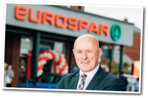 A man in a suit and tie poses in front of a EuroSpar store. Description generated by AI