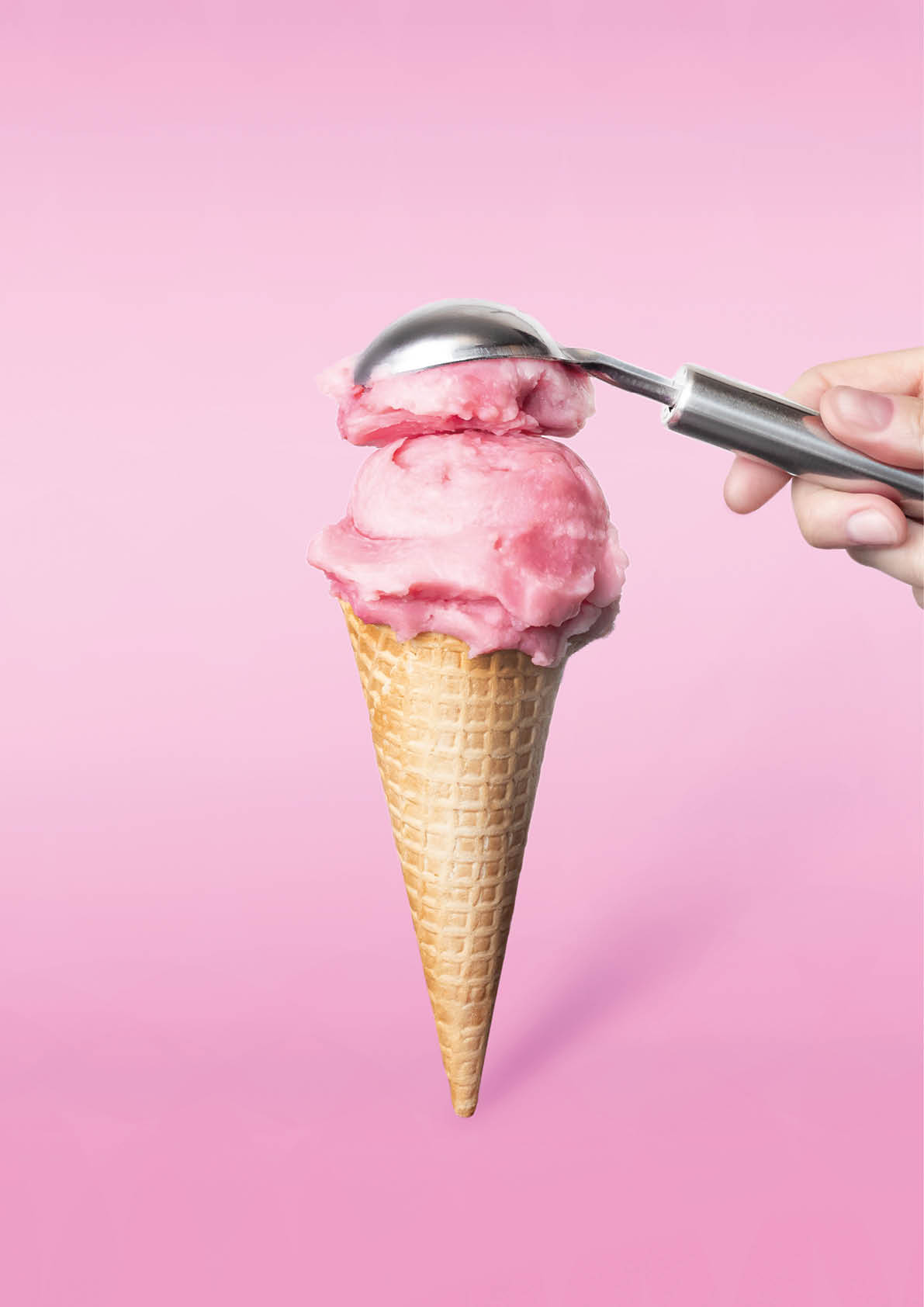 Strawberry ice cream waffle cone on pink background vertical food photo minimalism. Woman's hand puts a scoop of strawberry ice cream
