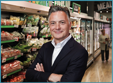 A person with short, styled hair wearing a dark blazer and white shirt stands with arms crossed and smiles in a grocery store aisle filled with fresh produce. Description generated by AI