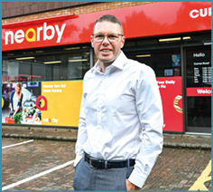 A person wearing glasses, a white button-up shirt, and gray pants stands smiling in front of a storefront with red and yellow signage that reads “nearby." Description generated by AI