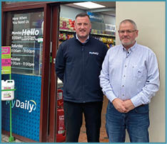 Two adults stand side by side outside a store with a glass door displaying business hours and promotional signs. Description generated by AI