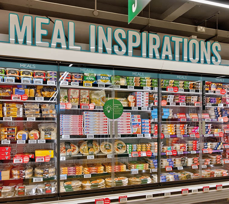A supermarket refrigerated section labeled “Meal Inspirations" displays a variety of packaged ready-to-eat meals and food items organized on shelves behind glass doors. Description generated by AI
