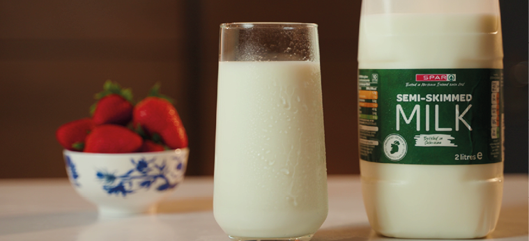 A glass of semi-skimmed milk with condensation sits on a table next to a bottle of milk and a bowl of strawberries in the background. Description generated by AI