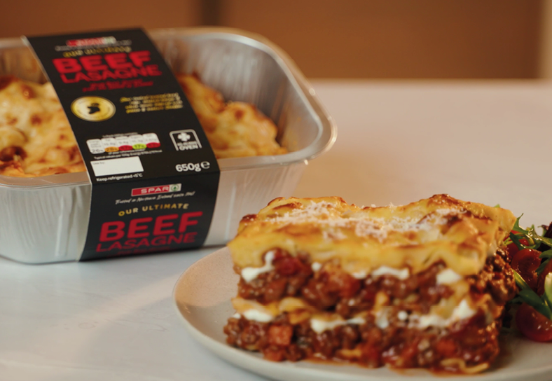 A serving of layered beef lasagne on a plate with a side of salad is placed in front of a foil container labeled as beef lasagne. Description generated by AI