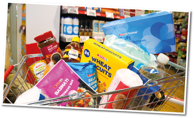 A shopping cart filled with various grocery items including wheat biscuits, tissue box, honey, spices, and packaged food products in a supermarket aisle. Description generated by AI