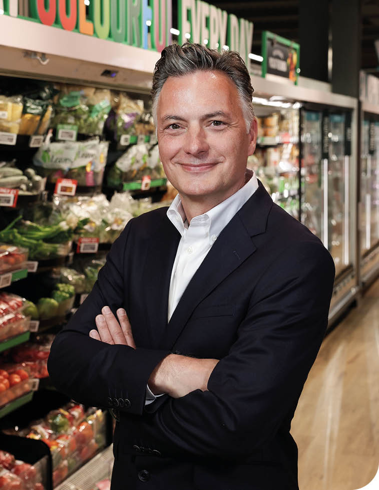 A person with short styled hair wearing a dark blazer and white shirt stands with arms crossed and smiles in a grocery store aisle filled with fresh produce. Description generated by AI