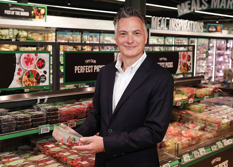 A person wearing a dark suit and white shirt is smiling while holding a container of strawberries in a grocery store produce section. Description generated by AI