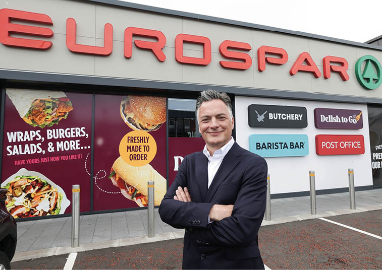 A person in a dark suit stands with arms crossed smiling in front of a Eurospar store with signs advertising wraps, burgers, salads, a butchery, barista bar, post office, and a deli. Description generated by AI