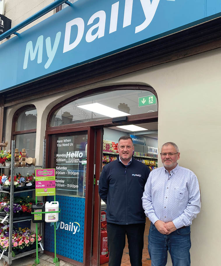 Two adults stand smiling in front of a convenience store entrance with a blue sign above reading “MyDaily" and shelves of flowers visible to the side. Description generated by AI