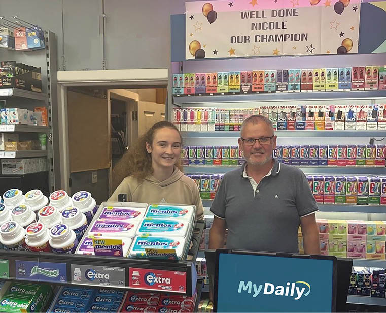 Two adults stand behind a counter in a store with shelves of colorful products and a congratulatory sign that reads “Well done Nicole our champion" above them. Description generated by AI