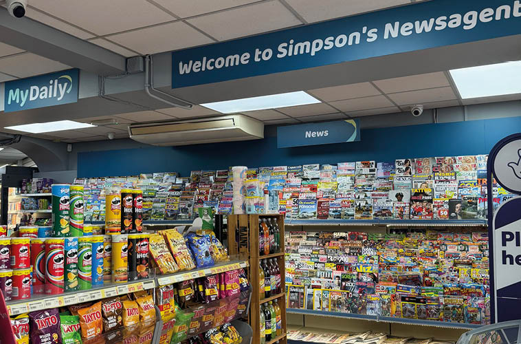 A brightly lit newsagent store interior with shelves stocked with colorful snack packages and a wall filled with various magazines and newspapers under signs reading Welcome to Simpson's Newsagent and MyDaily. Description generated by AI