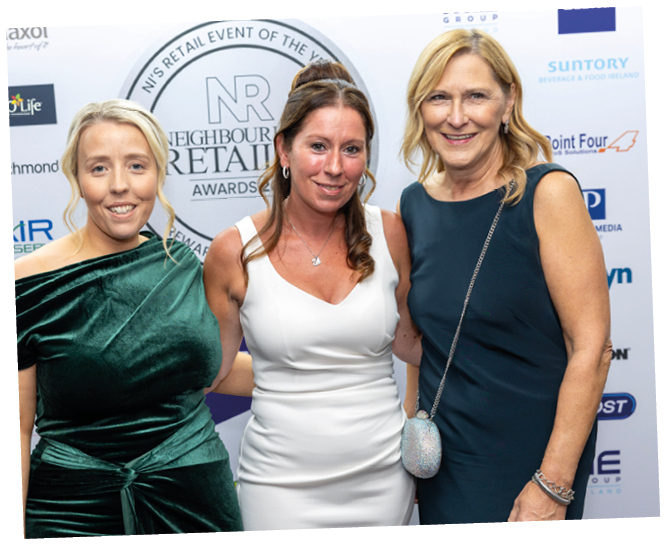 Three adults dressed in formal attire stand closely together smiling at a retail awards event with a branded backdrop behind them. Description generated by AI
