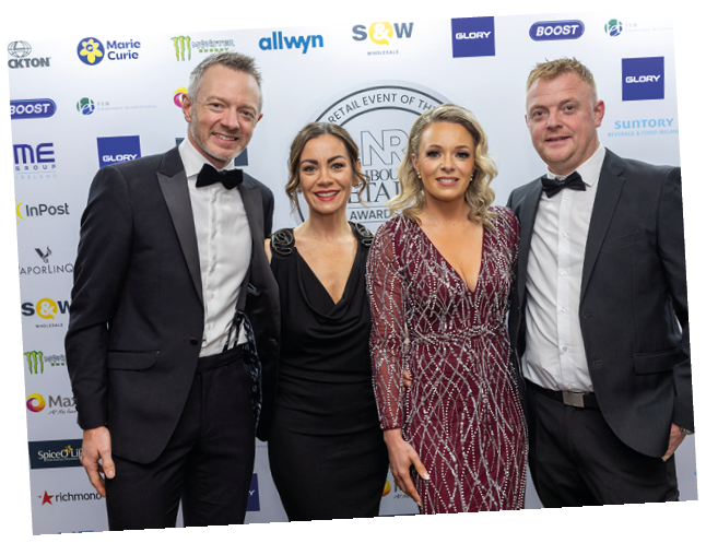 Four adults dressed in formal attire stand closely together smiling in front of a backdrop with various logos at an event. Description generated by AI