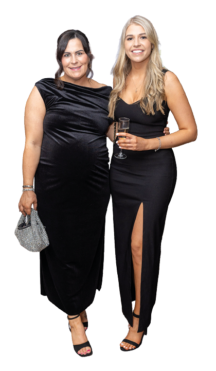 Two adults dressed in elegant black evening gowns stand closely together smiling, with one holding a small handbag and the other holding a glass of champagne. Description generated by AI