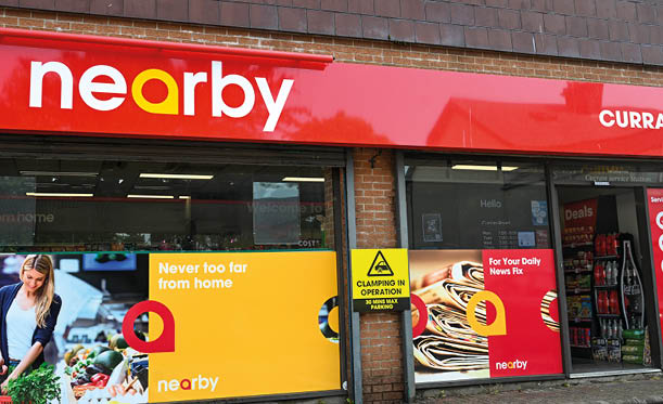 A storefront with a red sign reading “nearby" above windows displaying colorful advertisements for local news and products with a person arranging items on the left side. Description generated by AI