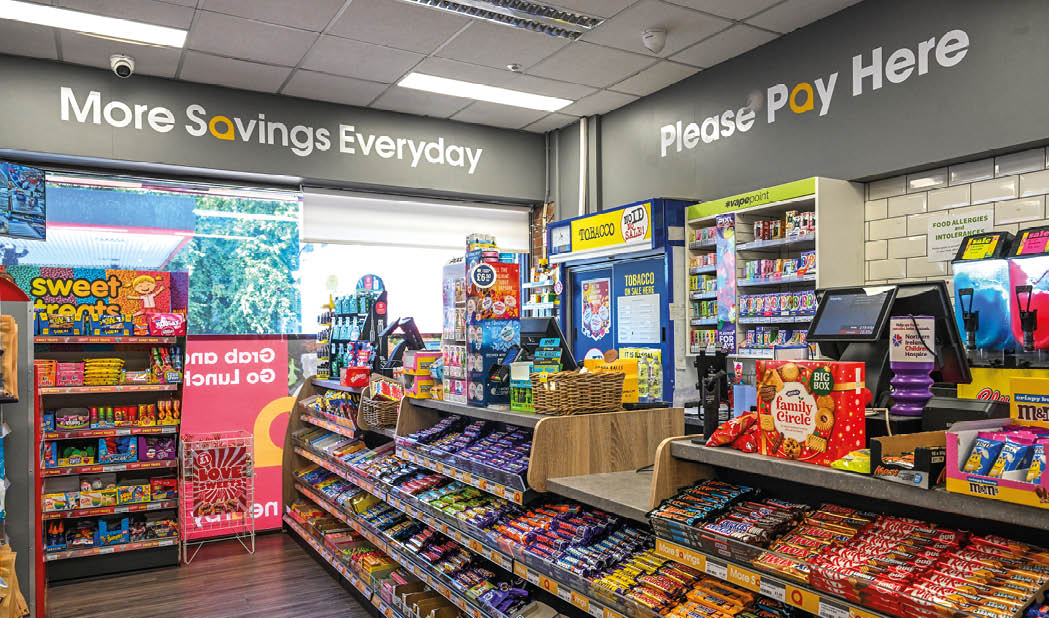 A brightly lit convenience store interior with shelves stocked with snacks, candy, and beverages, featuring signs that say “More Savings Everyday" and "Please Pay Here." Description generated by AI