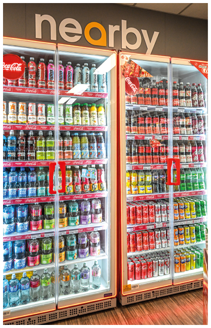 A brightly lit refrigerated display case filled with various colorful canned and bottled beverages arranged neatly on shelves under a sign that reads “nearby." Description generated by AI