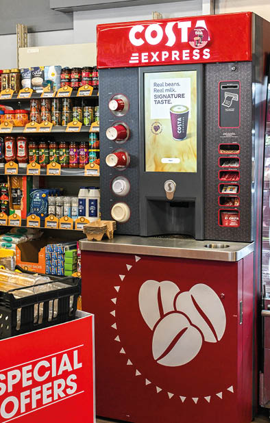 A Costa Express coffee vending machine stands next to shelves stocked with various grocery items and a sign advertising special offers. Description generated by AI