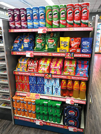 A supermarket shelf displays various snack products including multiple flavors of Pringles, Walkers crisps, M&M's, Maltesers, Skittles, Lucozade drinks, Fanta cans, Monster energy drinks, and Pepsi bottles, all labeled with special offer tags. Description generated by AI