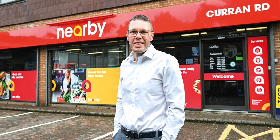 A person wearing glasses and a white button-up shirt stands smiling in front of a storefront with a red sign that reads “nearby" and "CURRAN RD." Description generated by AI