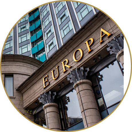 A building facade with large columns and the word EUROPA in gold letters above the entrance is shown with a modern glass building in the background. Description generated by AI