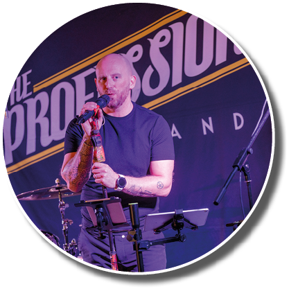 Person with tattoos wearing a black t-shirt is speaking into a microphone on a stage with musical instruments and a backdrop that reads “The Profession." Description generated by AI